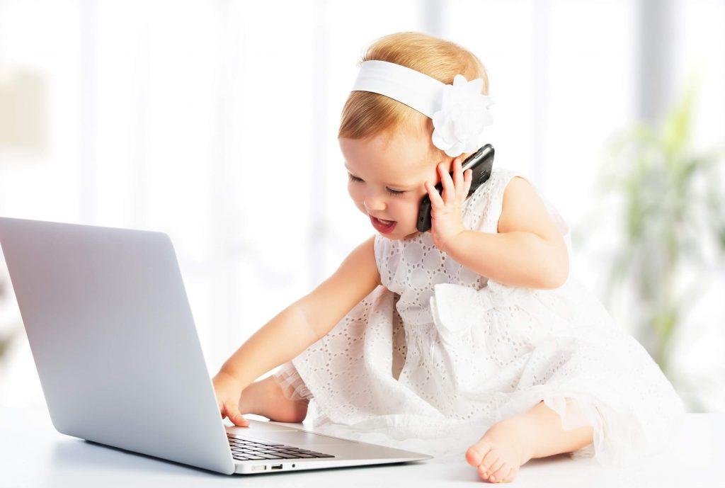 Ein Baby gekleidet mit einem weißen Kleid und einem Haarband sitzt vor einem Computer und telefoniert dabei mit einem Handy